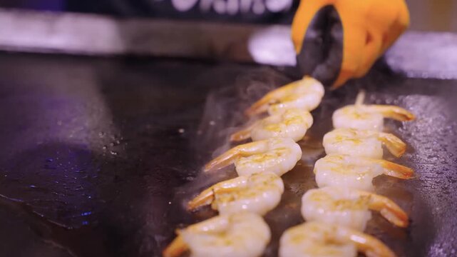 High-angle close-up of fresh shrimp skewers being cooked with a professional culinary blowtorch on a hot griddle. The footage captures the intense blue flame searing the succulent pink shrimp, creatin