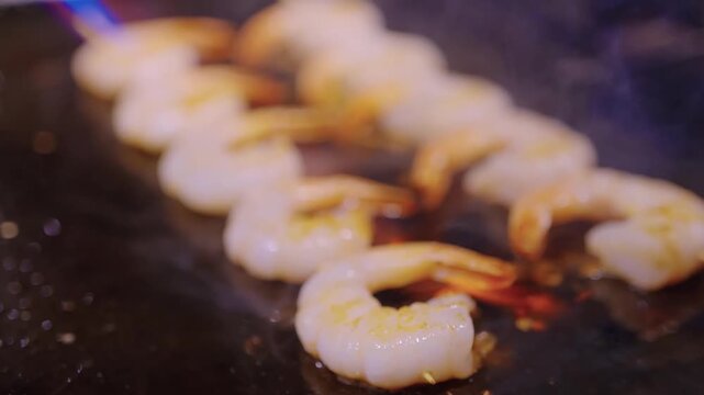 Professional culinary footage showcasing a row of fresh shrimp skewers being seared with an open flame blowtorch on a hot metal griddle. The high-heat cooking process creates a perfect char and smoky 
