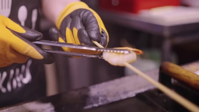 Close-up footage of a professional chef using metal tongs to flip and sear succulent Hokkaido scallops and jumbo shrimp on a hot teppanyaki flat-top grill. The video captures the steam and golden-brow