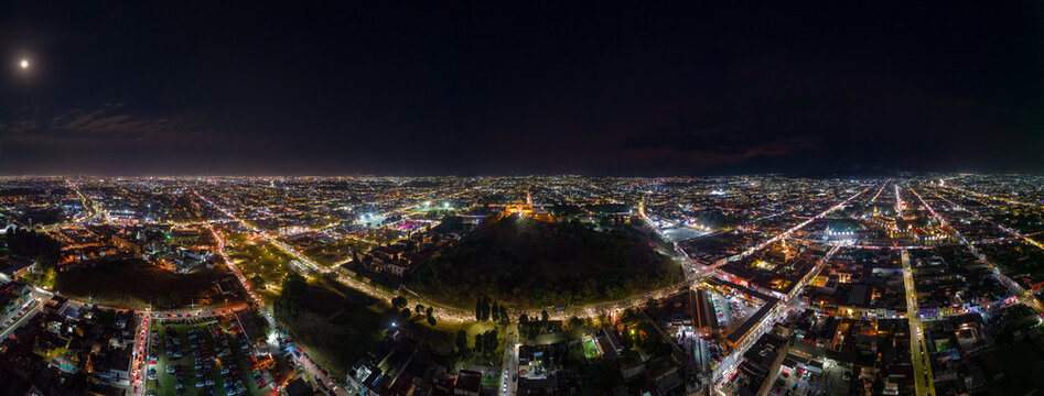 San Pedro Cholula, Puebla, Mexico - 05 March 2023: Aerial view of the Great Pyramid of Cholula illuminated at night, surrounded by the glowing city lights and urban grid.