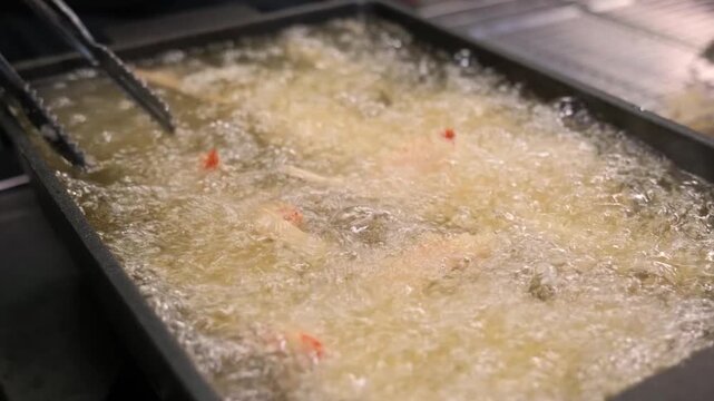 Close-up footage of traditional Japanese tempura being deep-fried in a professional kitchen. The chef uses long tongs to manage shrimp and vegetables in bubbling hot oil, achieving a light, golden-bro