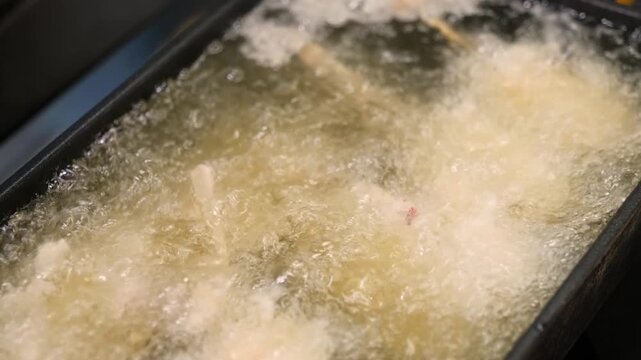 A high-angle, close-up shot capturing the intense cooking process of food&mdash;likely fish, shrimp, or chicken&mdash;being deep-fried in a professional stainless steel fryer. The hot oil is bubbling vigorously a