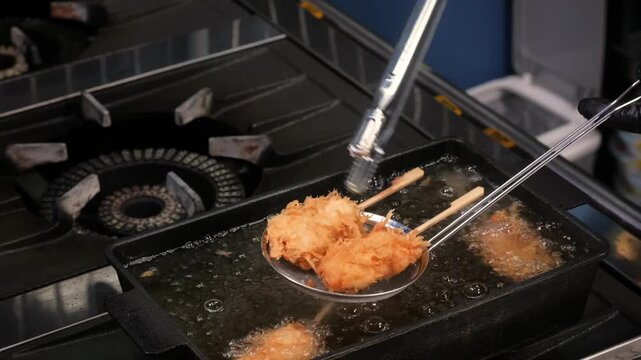 A close-up view of a professional chef using tongs to carefully deep fry traditional Japanese Kushiage (kushikatsu) skewers in a rectangular pan with hot, bubbling oil within a restaurant kitchen envi