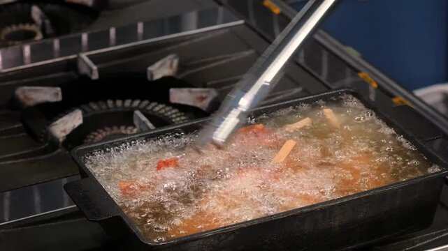 Professional kitchen footage showing breaded chicken pieces being deep-fried in hot, bubbling vegetable oil until golden brown. A chef uses metal tongs to carefully handle the crispy chicken. Ideal fo