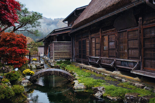 Shirakawa, Gifu, Japan - 09 November 2025: View of Shirakawa-go featuring a traditional thatched-roof house, a small stone bridge over a stream, and vibrant autumn foliage.