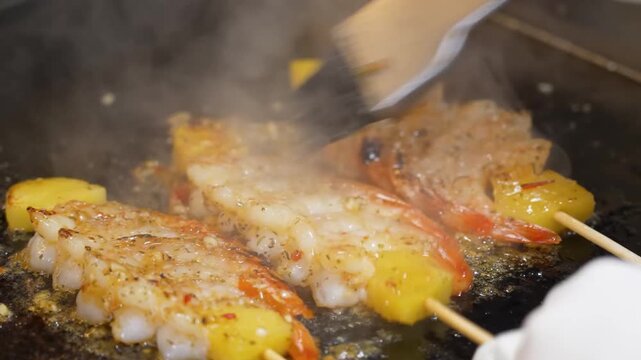 close-up, high-angle shot capturing a professional chef using a metal spatula to stir and toss fresh shrimp and pineapple chunks on a sizzling hot commercial griddle. The footage highlights the vibran
