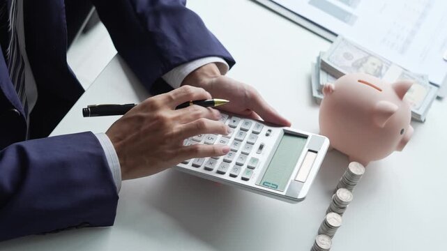 Businessman in suit pressing calculator with biggy bank and stacking coins