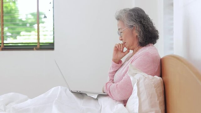 Happy elderly woman wearing glasses sitting in bed and typing on laptop, freelancing or staying connected with family from home.