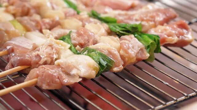 Close-up footage of a professional chef's hands in gloves preparing and arranging raw chicken and green onion (negima) yakitori skewers on a traditional grill. This authentic Japanese street food and 