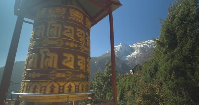 Namche Bazaar, Nepal, static low angle shot of a large spinning Buddhist prayer wheel engraved with mantras against the backdrop of snow-capped Himalayan mountains.