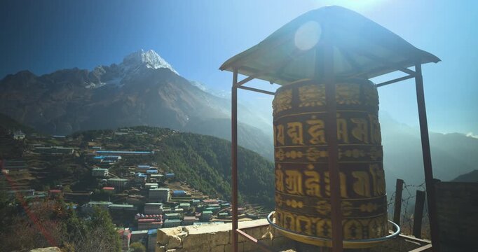 Namche Bazaar, Nepal, static low-angle shot of a large spinning Buddhist prayer wheel overlooking colorful rooftop houses and Kongde Ri mountain peak under bright sunlight.