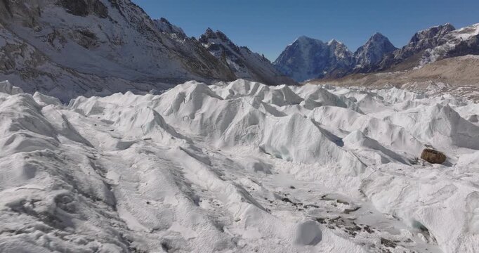 Khumbu Glacier, Everest Nepal, drone aerial shot moving smoothly over a field of white ice waves and jagged seracs with rugged mountain peaks rising in the distant background under a clear blue sky.