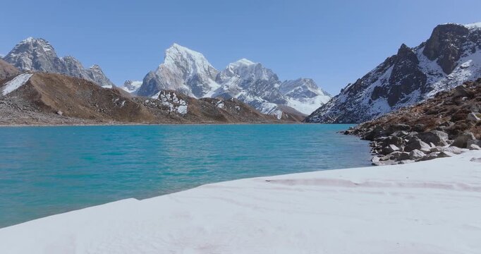 Aerial view gliding across the pristine blue waters of Gokyo Lake with majestic Himalayan mountains under bright sunny skies in the Everest Region, Nepal.