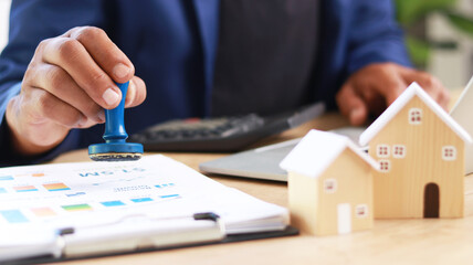 Businessman stamping approval on real estate documents with house models on desk, symbolizing...