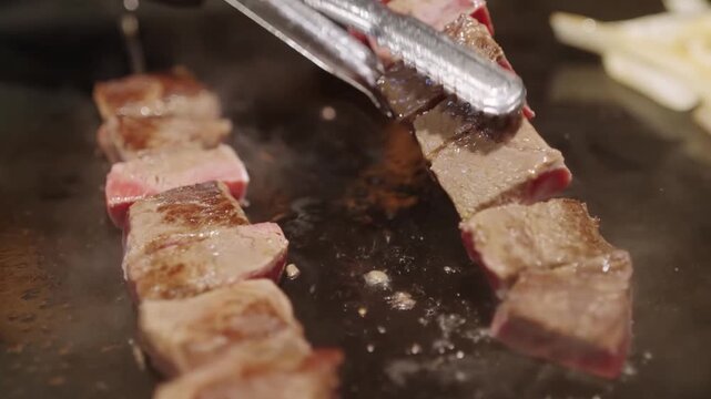 Close-up of premium A5 Japanese Wagyu beef cubes and slices sizzling on a hot teppanyaki grill. The footage highlights the exquisite fat marbling as the meat cooks, releasing steam and rich juices in 