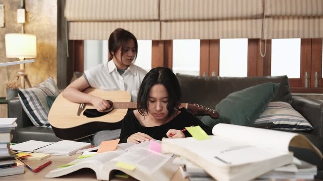 Two Asian teenage tutorials, girl concentrates on reading books for exam, learning at home, her friend relaxes by playing guitar music, self-study with textbooks, homeschooling together lifestyle.