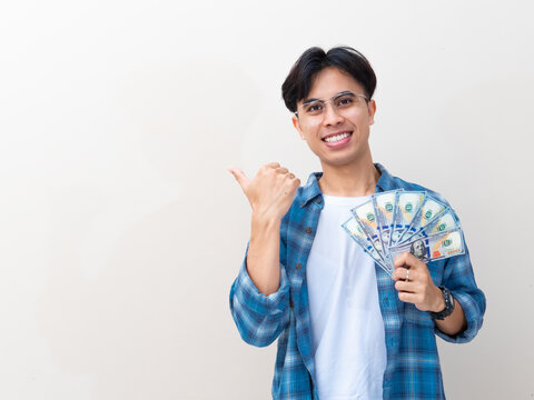 A happy young Asian man smiling at the camera while holding a fan of cash and pointing sideways with his thumb against a plain studio background.