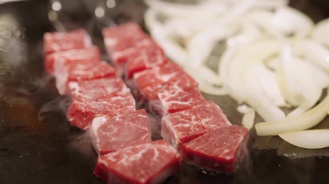 A high-angle, close-up shot of premium marbled beef cubes (wagyu or high-quality sirloin) and sliced onions sizzling on a flat-top grill. The footage captures the steam and initial searing process typ