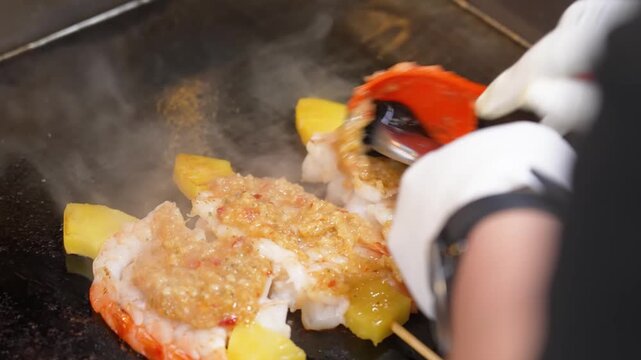 High-angle shot of a chef expertly preparing lobster tails on a flat-top teppanyaki grill. The chef, wearing white gloves, is seen basting the succulent lobster meat with a rich, golden garlic and gin