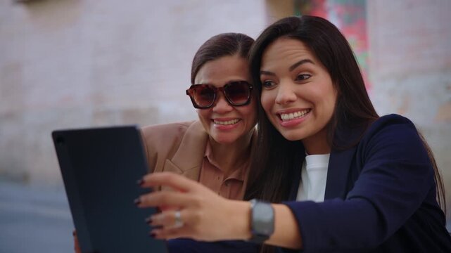 Two multi-generational businesswomen hold a digital tablet and smile as they prepare to take a selfie together on an urban street.