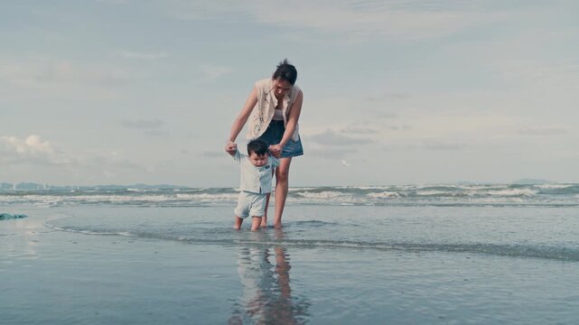 4K Video of an Asian mother holding her baby boy's hands, supporting his first steps on the beach.