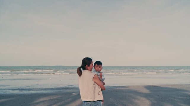 4K Video of an Asian mother holding her baby boy and looking at the sea view on the beach.