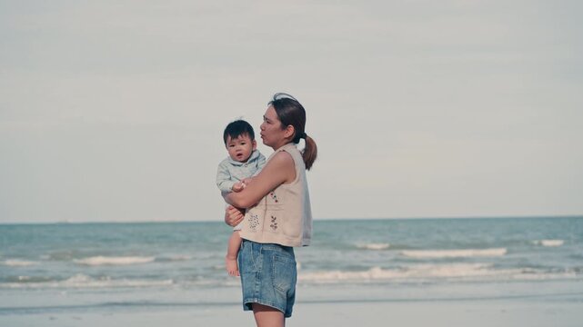 4K Video of an Asian mother holding her baby boy and looking at the sea view on the beach.