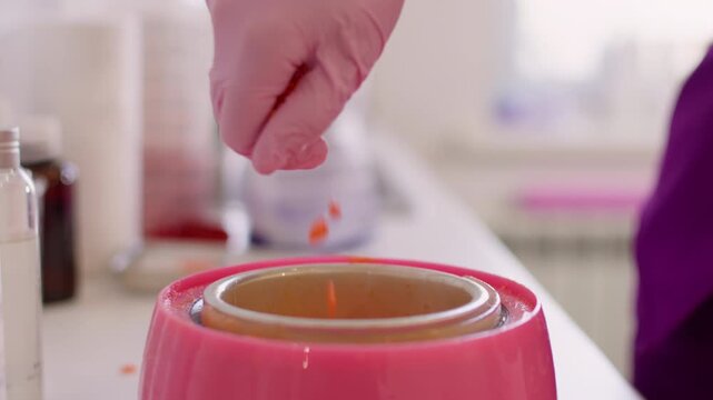 Wax pellets melting . Body care. Female hand in pink latex glove using orange product granules in heater preparation for professional cosmetic procedure.