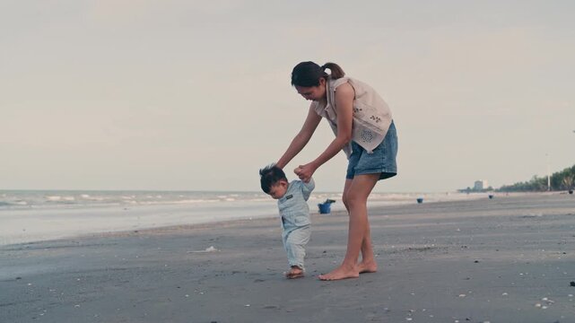 4K Video of an Asian mother holding her baby boy's hands, supporting his first steps on the beach.