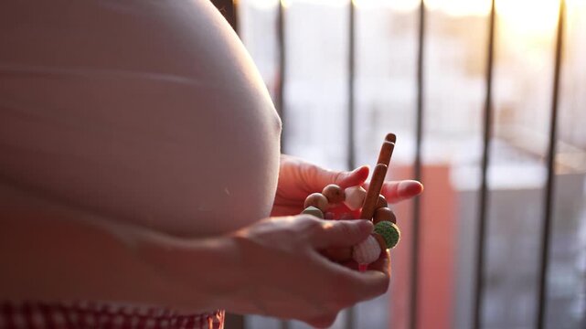 Expectant mother holding a small wooden rocking horse teether in her hands near her pregnant belly, standing on a balcony during a beautiful, warm sunset, embodying preparation for childbirth