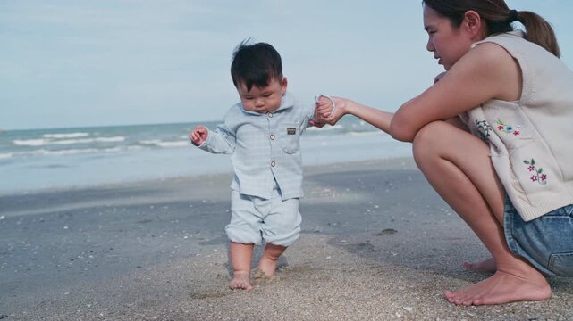 4K Video of an Asian mother holding her baby boy's hands, supporting his first steps on the beach.