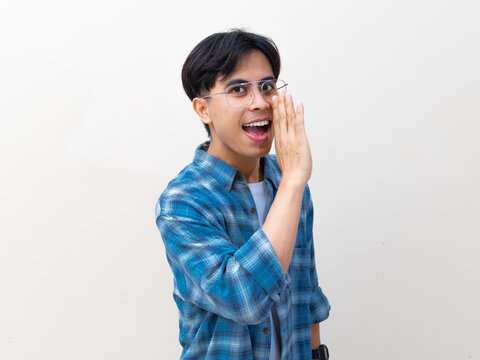 A friendly young Asian man looking at the camera while whispering a secret or making an announcement against a plain studio background.