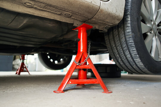 Closeup of red jack stand supporting the underbody frame of a lifted car during auto repair or maintenance work