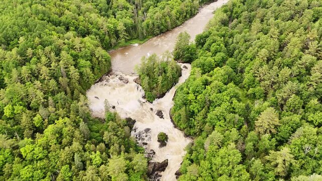 Drone gradually tilts down revealing top of Chutes de Plaisance, Quebec. Spring high water, cascading falls, lush green forest, aerial overhead perspective.