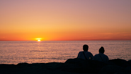 Fototapeta premium Silhouette of a couple sitting on rocks and watching the sunset over the sea.