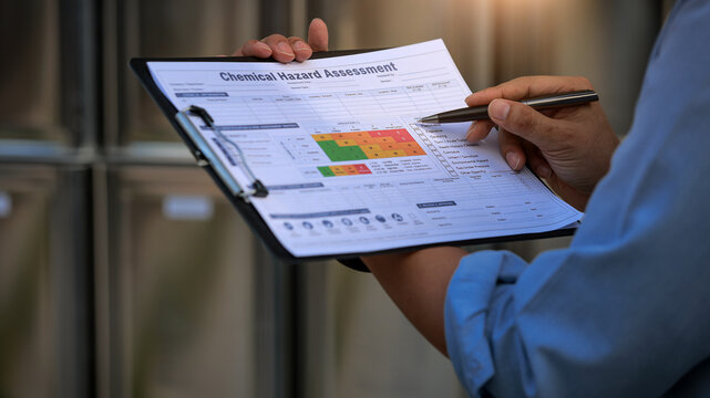 Focused inspector reviews chemical hazard assessment document on clipboard ensuring safety storage compliance inside industrial warehouse facility