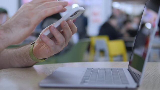 Hands tap phone while laptop sits nearby. Person focused on mobile device. Background shows public space with chairs and people. Suitable for tech, work, or multitasking concepts