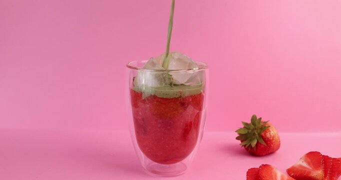 Pouring green matcha milk over strawberry puree layer in glass on pink background. Layered iced beverage concept features cold tea cocktail with ice cubes and fresh red berries. Colorful contrast shot