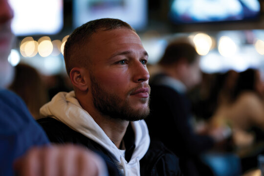 A moment of calm captures a young man with a beard, as he appears lost in thought, surrounded by a busy scene, reflecting the deep contemplation amidst social interaction.