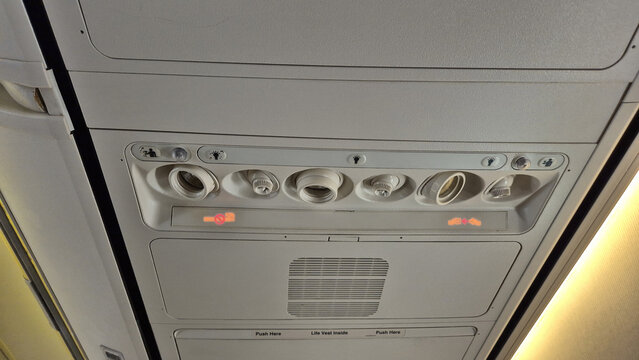 An overhead passenger service unit inside an airplane displays reading lights, air vents, a illuminated no-smoking symbol, and seatbelt signs.