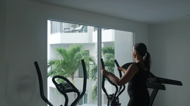Young woman in black sportswear using an elliptical machine in a modern gym with large glass windows overlooking tropical palms and a pool, slow motion. Fitness concept