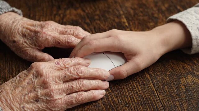 Wrinkled elderly hand and young hand touching computer mouse wooden table symbolizing connection support care technology use and gentle focused mood