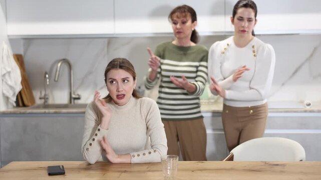 Sad middle-aged woman sitting at table and other women brawling to her standing behind in the kitchen