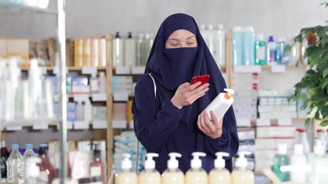 Young female customer dressed in traditional Muslim attire, dark abaya and khimar(niqab), holding body lotion bottle(tube, package) and smartphone, comparing ingredients or prices in drugstore