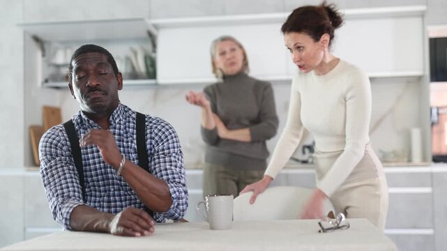 Upset middle-aged man sitting at table while two female family members quarreling to him standing behind in the kitchen. High quality 4k footage