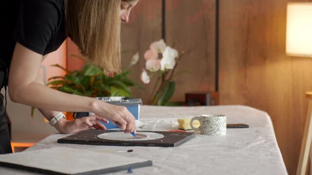 Woman applies white plaster paste to black canvas at a covered table in soft daylight. Concept of handmade wall decor abstract art and creative hobby