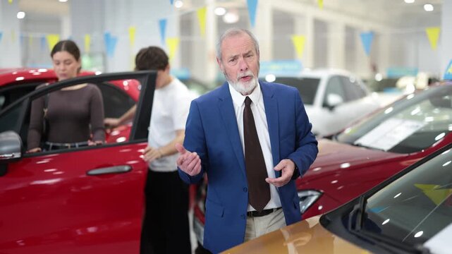 Elderly car salesman stands in car dealership and offers various cars to potential buyers