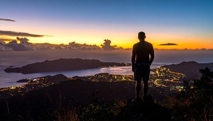 Fototapeta premium Silueta de hombre en la cima de una montaña contemplando una ciudad costera iluminada al atardecer