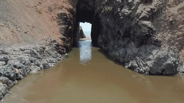 Tidal creek flows through narrow rock archway. Rocks frame murky water, showing nature's rugged beauty. Sunlight glints through the passage, casting gentle reflections. Ideal for travel, nature