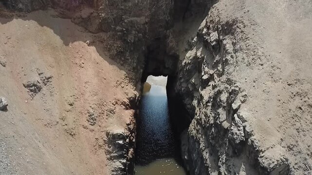 Rugged canyon frames a mysterious waterway. Sunlit rock walls frame a deep, dark passage. Water flows calmly through the narrow channel. Hidden stream invites curiosity and wonder